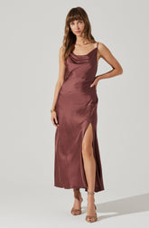 A woman is wearing a long, satin slip dress in a mauve color. The dress features thin spaghetti straps, a cowl neckline, and a high slit on the left side exposing her leg. She is also wearing open-toe high heels with ankle straps.